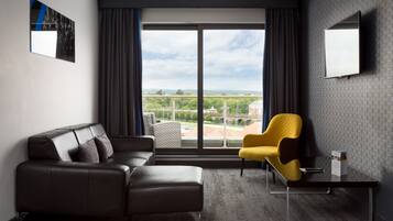 Une chambre d’hôtel moderne avec un canapé en cuir, un fauteuil jaune, un téléviseur à écran plat et une grande fenêtre donnant sur la ville.