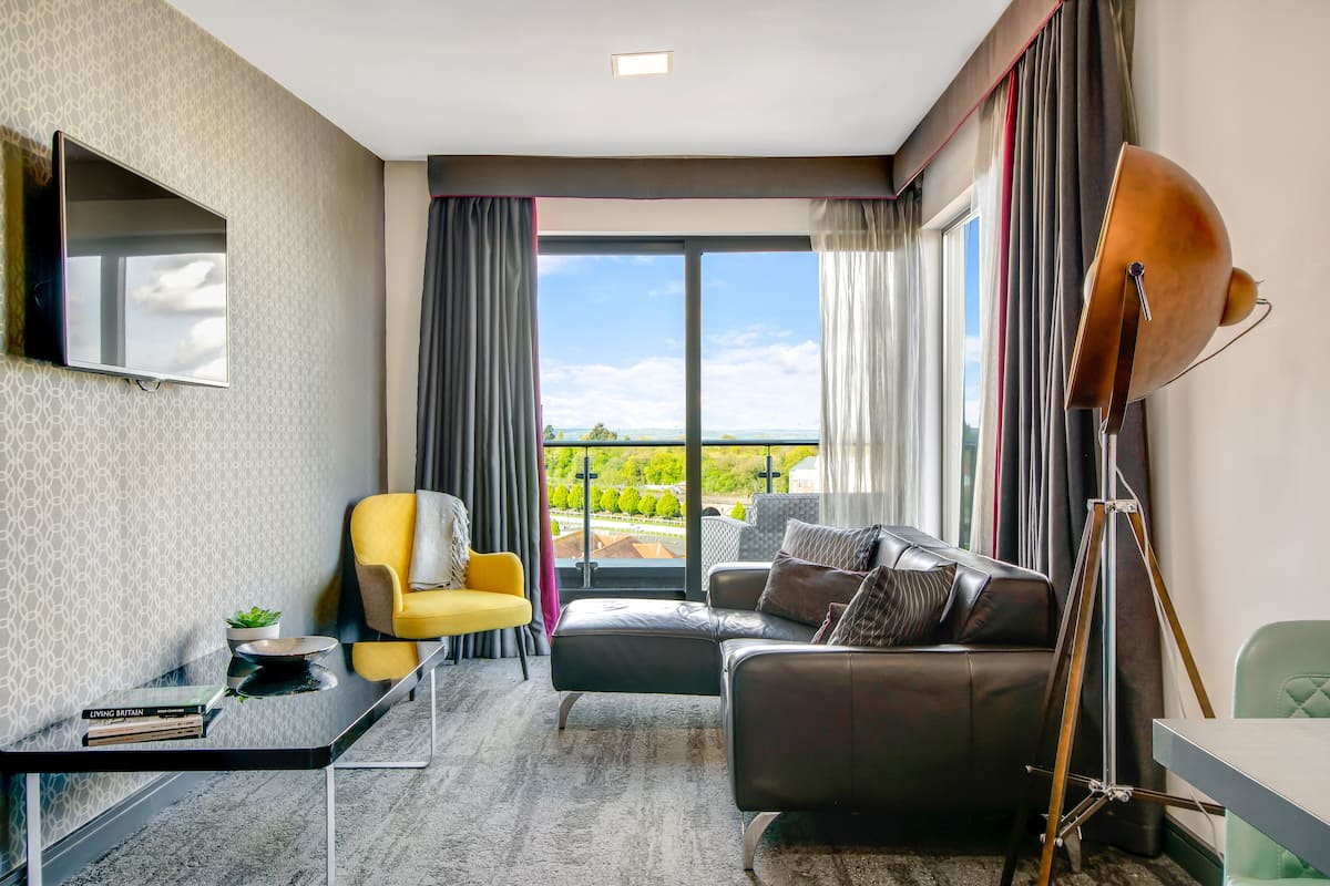 A modern living room with a large window, a flat-screen TV, a yellow armchair, a leather sofa, and a floor lamp.