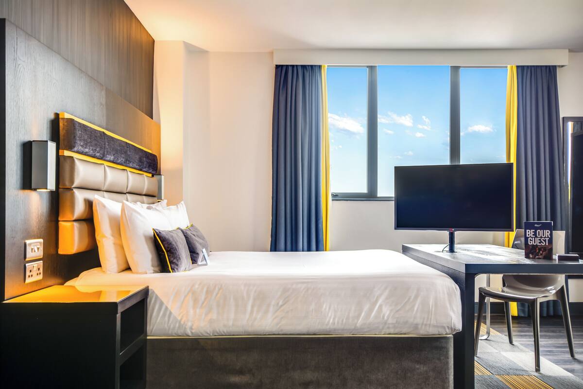 A modern hotel room with a large bed, a desk, a flat-screen TV, and a view of the sky through the window.