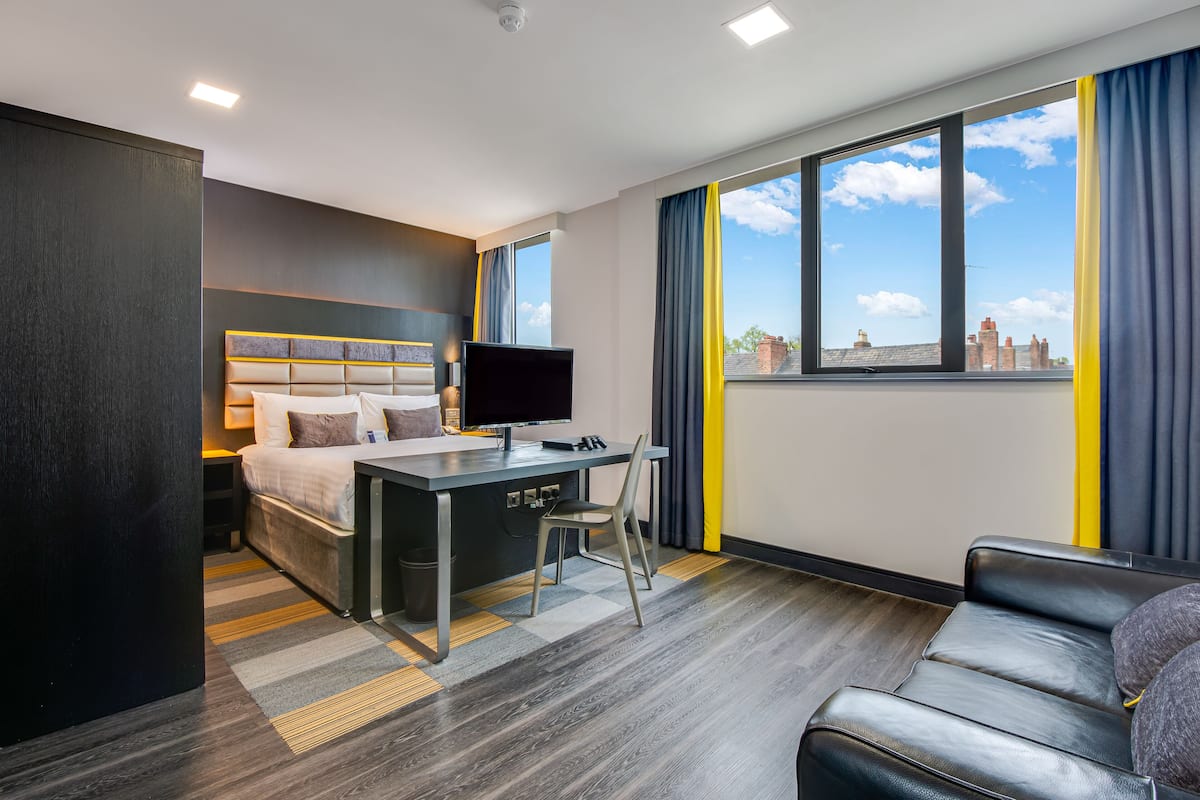 A modern hotel room with a bed, desk, chair, and a view of the cityscape through large windows.