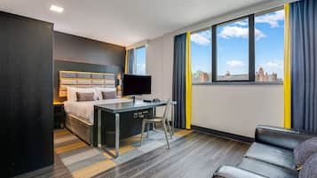A modern hotel room with a bed, desk, chair, and a view of the cityscape through large windows.
