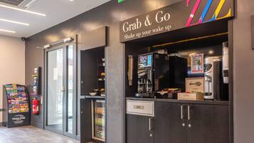 Una moderna zona de recepción de hotel con servicio 'Grab & Go', una pantalla digital, una máquina de café y una máquina expendedora.