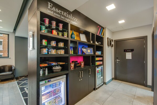 Un vestíbulo de hotel con un quiosco de autoservicio, un refrigerador con refrigerios y una puerta con la etiqueta 'UC'.