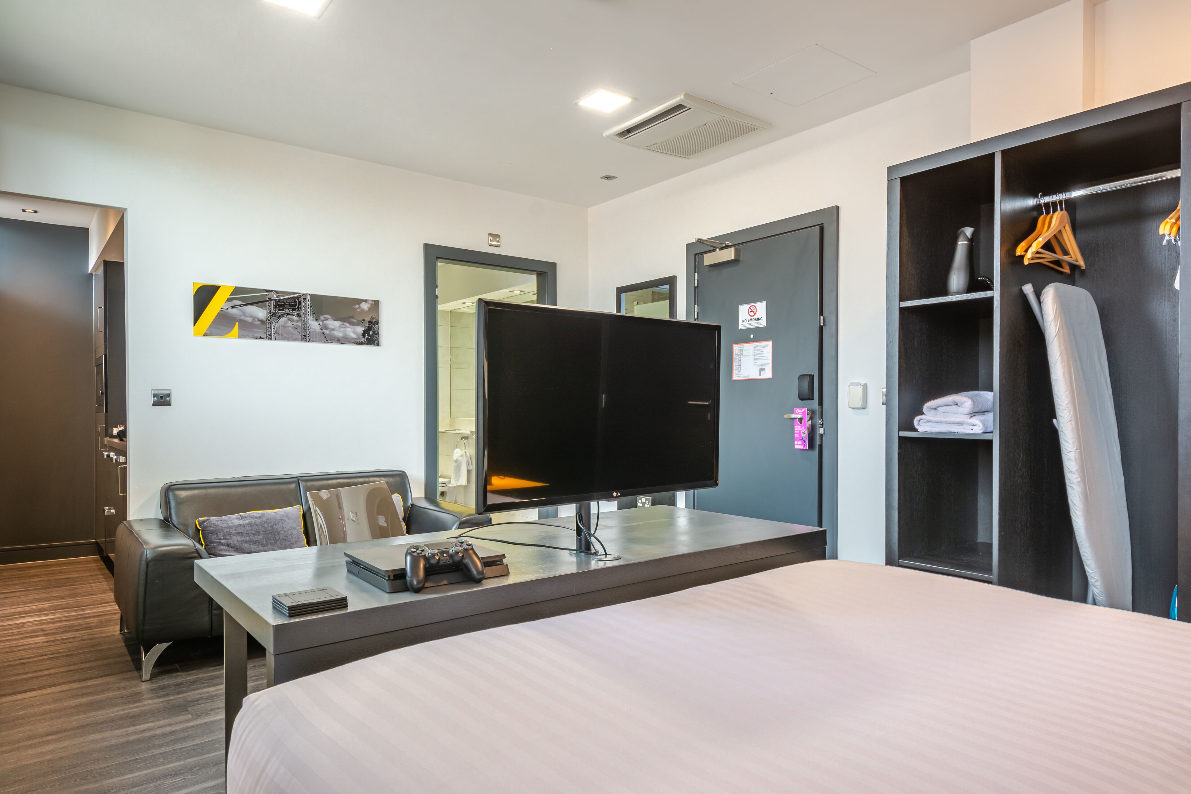 Una habitación de hotel con cama, escritorio, computadora, control de videojuegos, televisor, armario y perchero.