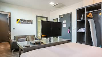 Una habitación de hotel con cama, escritorio, computadora, control de videojuegos, televisor, armario y perchero.