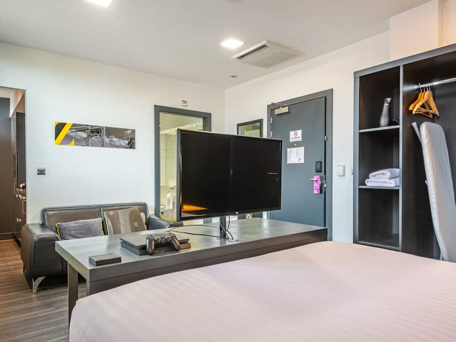 Una habitación de hotel con cama, escritorio, computadora, control de videojuegos, televisor, armario y perchero.