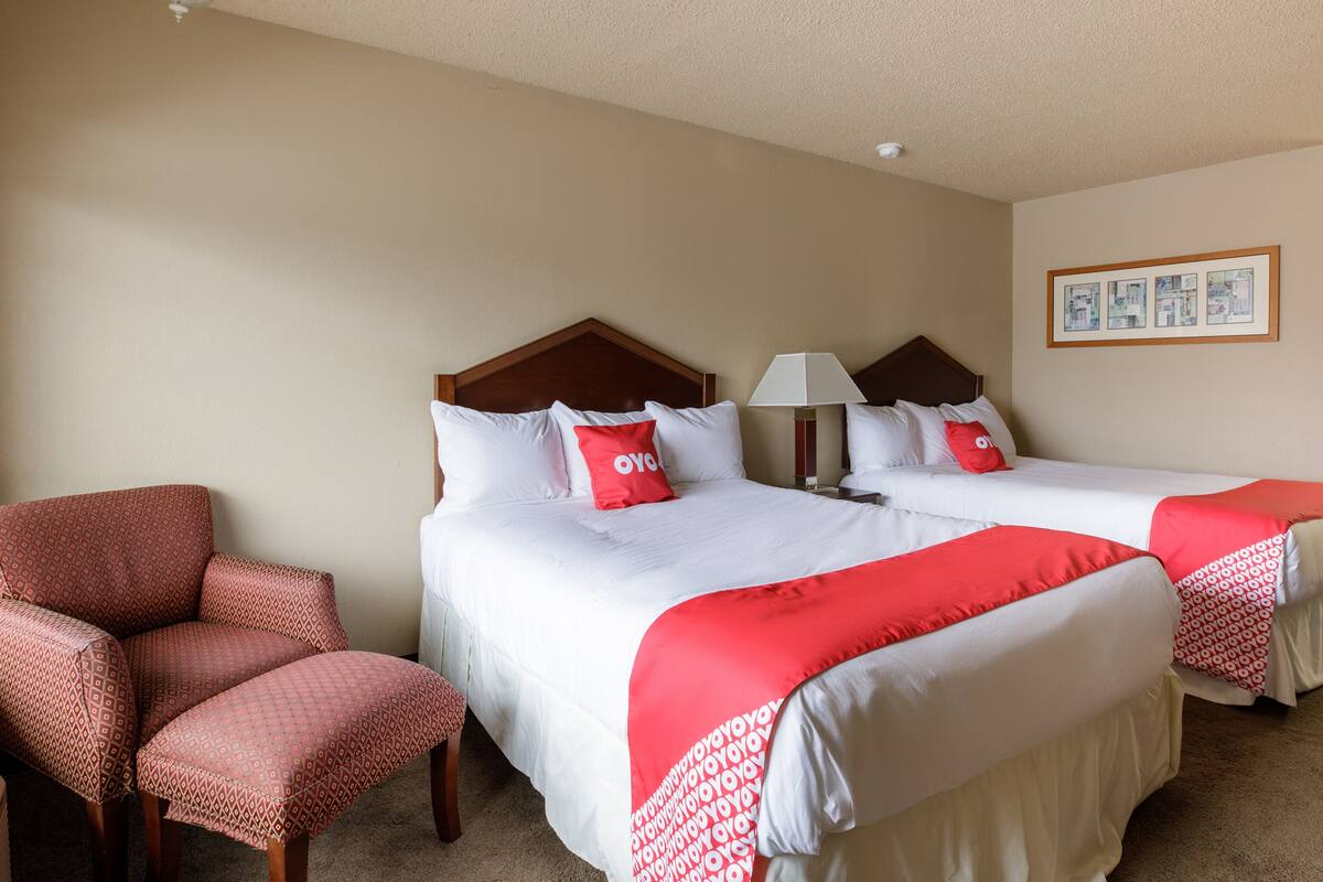 A hotel room with two beds, a chair, a lamp, and a ceiling light.