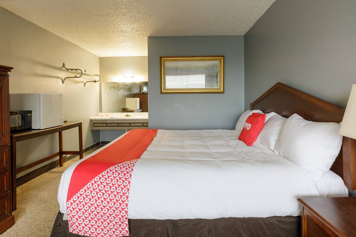 A hotel room with a bed, a bedside table, a nightstand, a desk with a microwave, a mirror, and a framed picture on the wall.