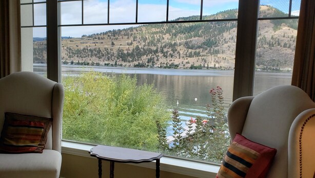 A room with a large window offering a view of a lake and hills. There are two chairs with pillows, a small table, and a bed with a white cover. - OKeefe's Landing Bed & Breakfast (Vernon)