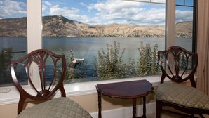 A room with two chairs and a small table by a window overlooking a lake with mountains in the background. - OKeefe's Landing Bed & Breakfast (Vernon)