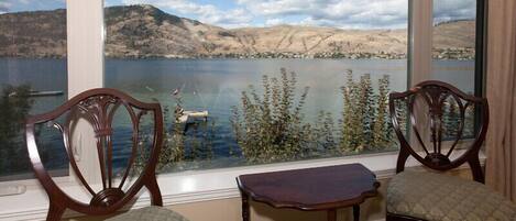 Une pièce avec deux chaises et une petite table près d’une fenêtre donnant sur un lac, avec des montagnes en arrière-plan.