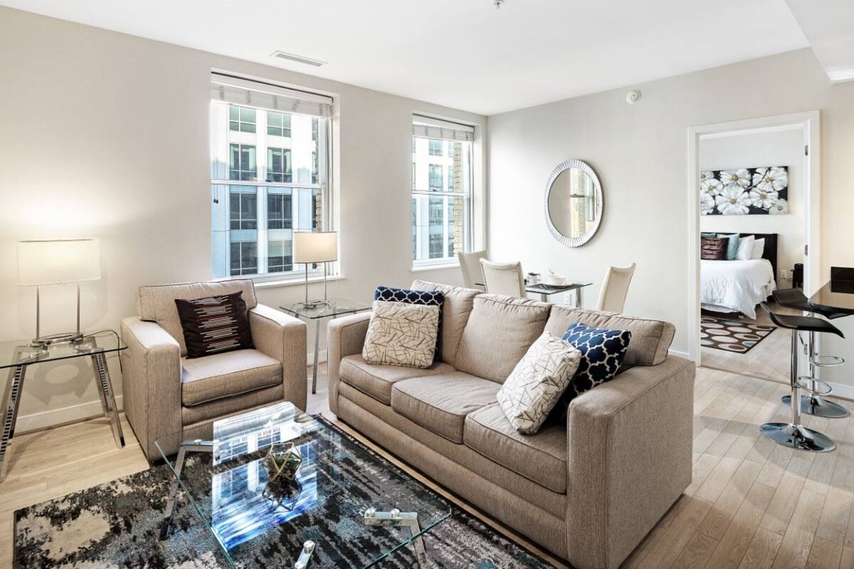 Ein modernes Wohnzimmer mit einem beigen Sofa, einem Glastisch und einem kleinen Beistelltisch. Zwei Fenster mit Jalousien, ein Spiegel an der Wand und eine Tür zu einem anderen Raum.