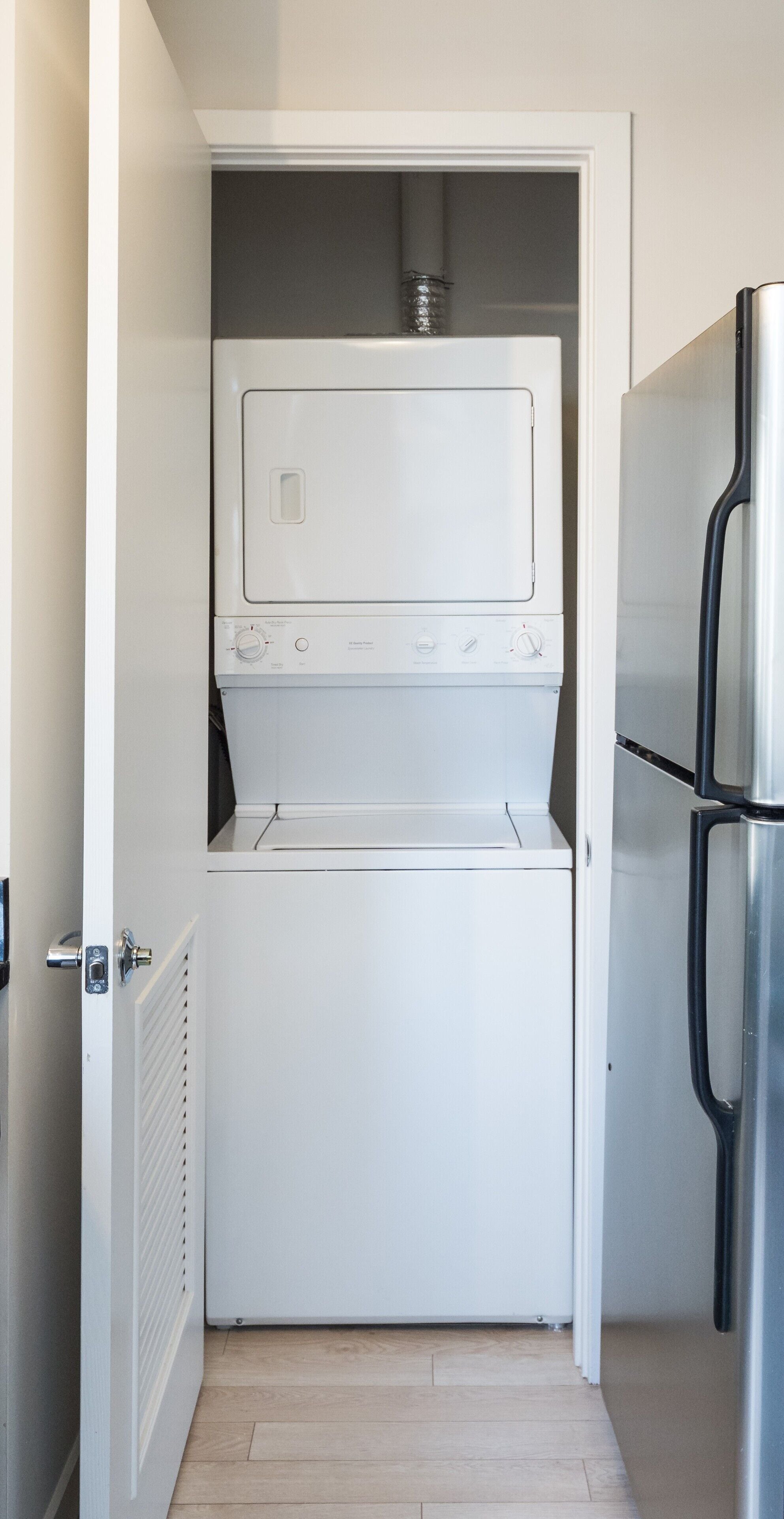 A compact laundry room with a white washer and dryer, a stainless steel refrigerator, and a wooden floor.