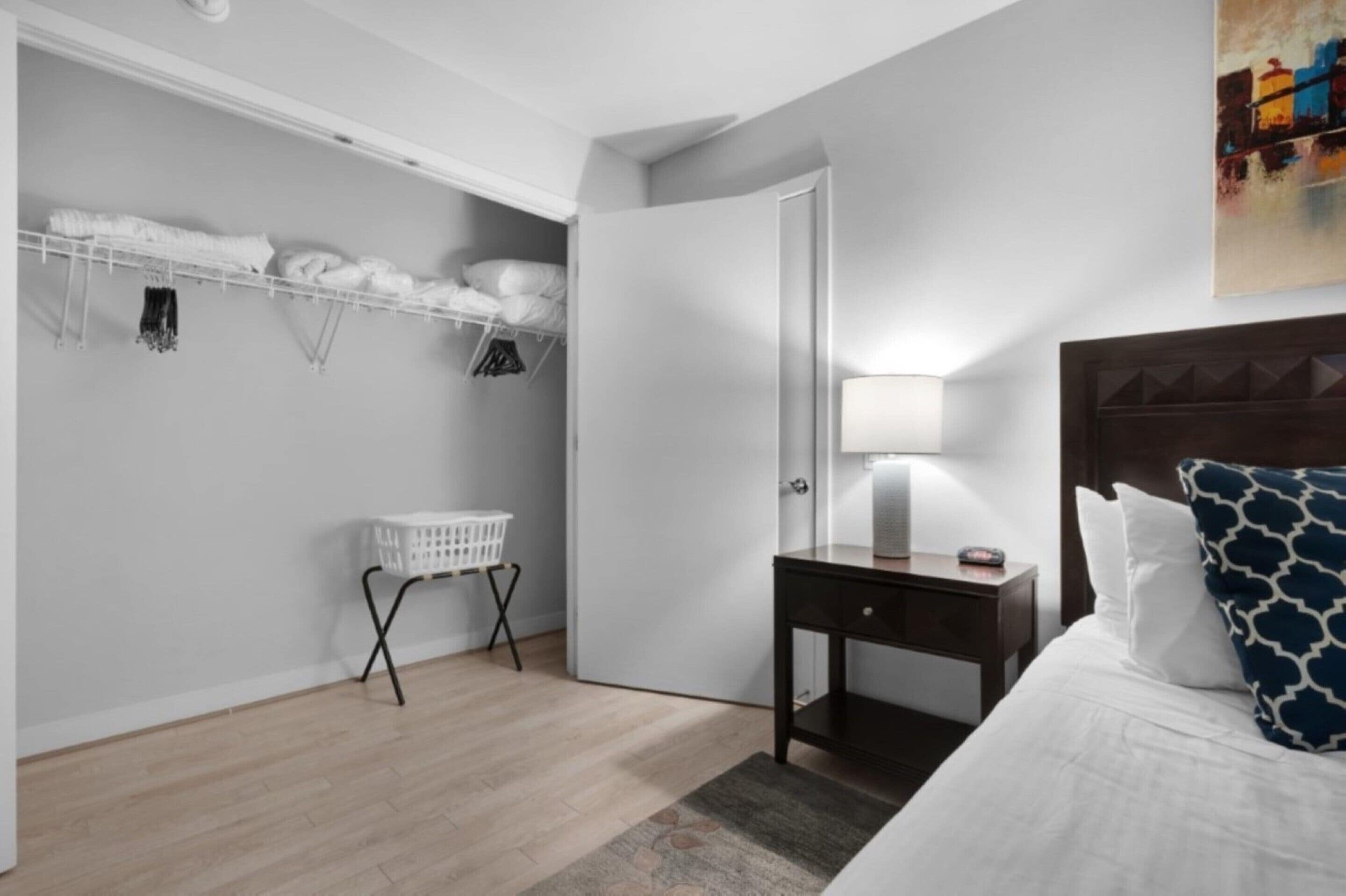 A hotel room with a bed, a nightstand with a lamp, a wardrobe with hanging clothes, and a laundry basket.