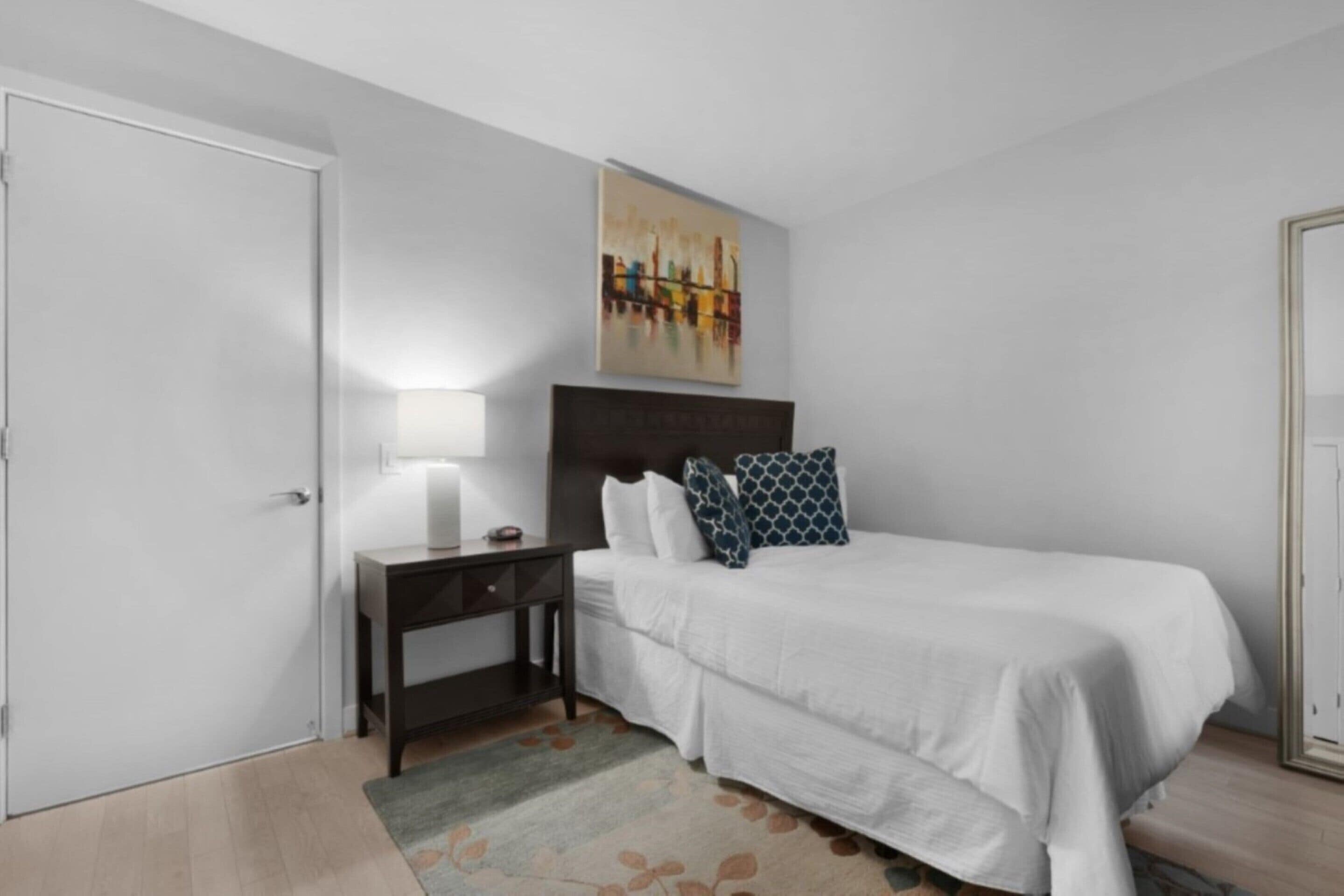 Un dormitorio con cama, mesita de noche, lámpara, un cuadro en la pared y una puerta.