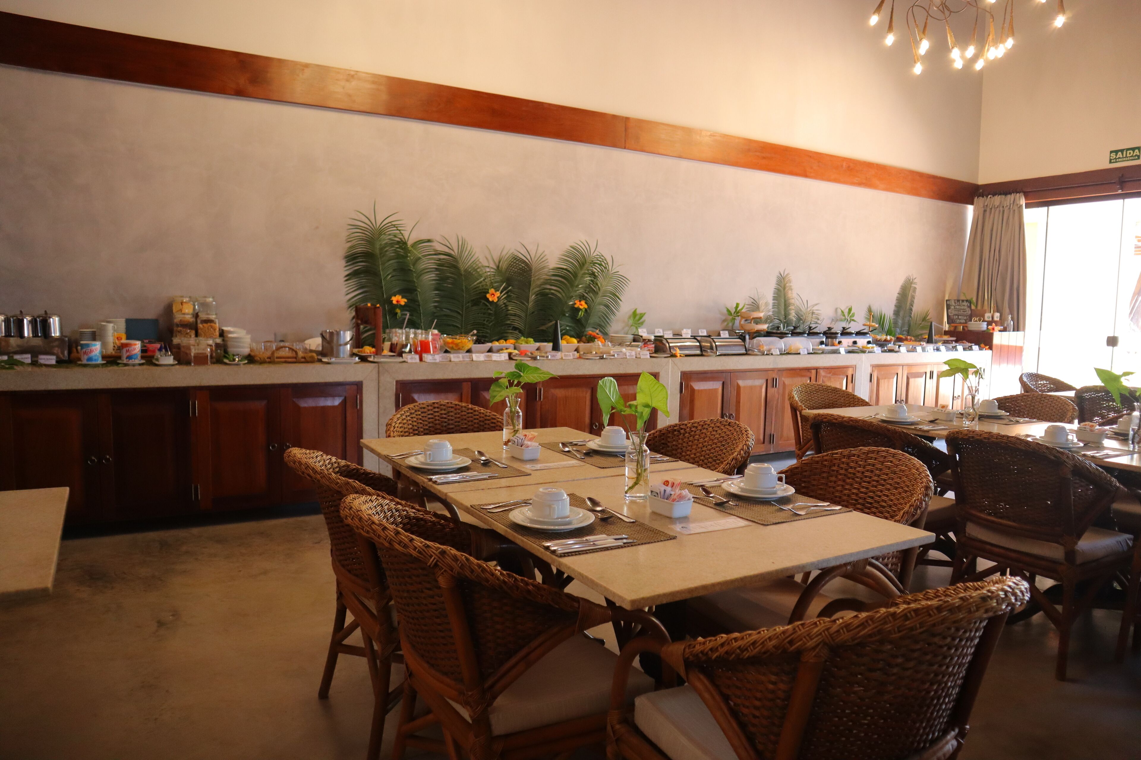 A dining area with wicker chairs, a long table set with plates, cups, and cutlery. There are potted plants and a buffet counter with various food items.