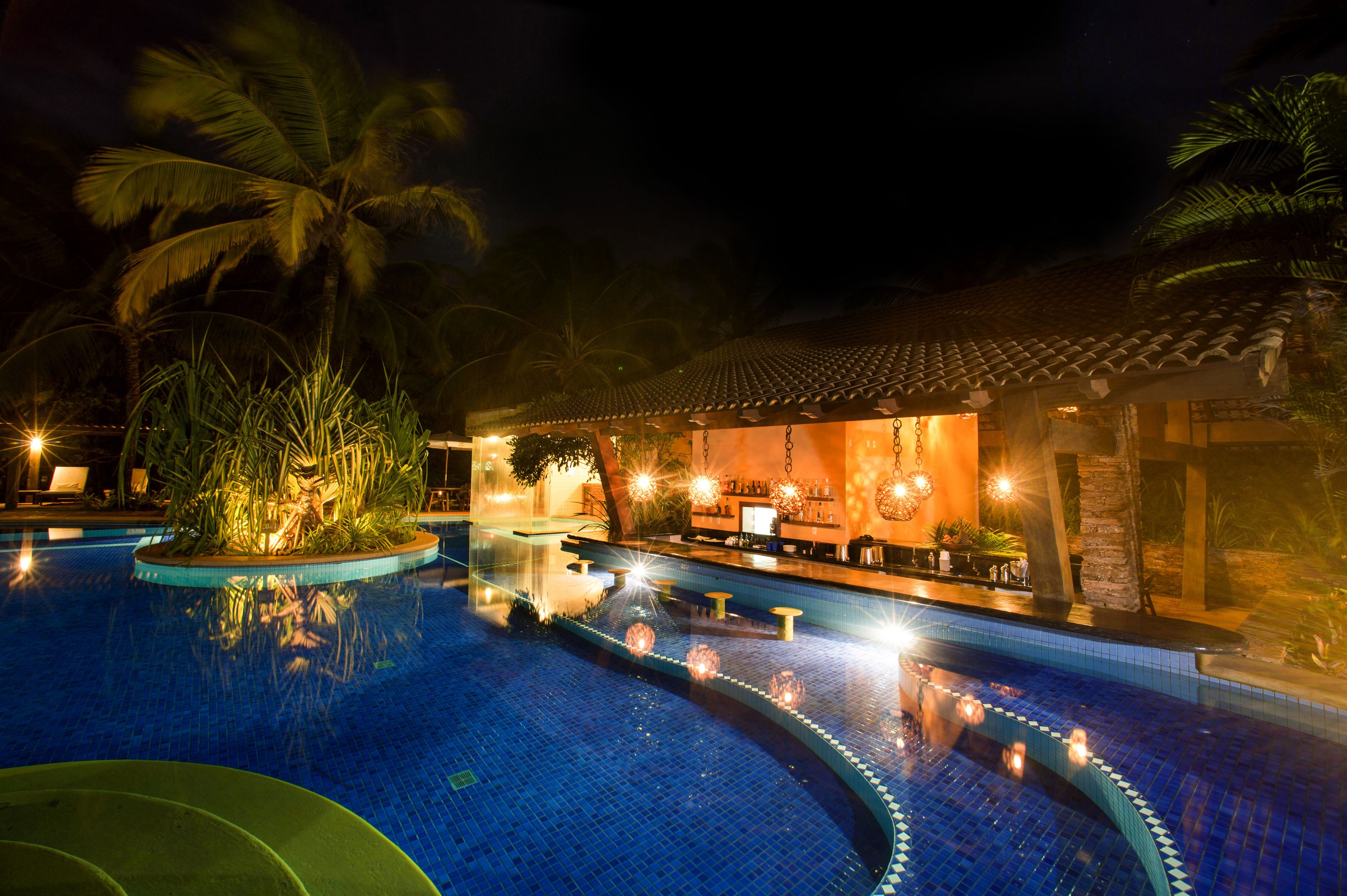 Zona de piscina nocturna con bar, área de descanso, palmeras y piscina con baldosas.
