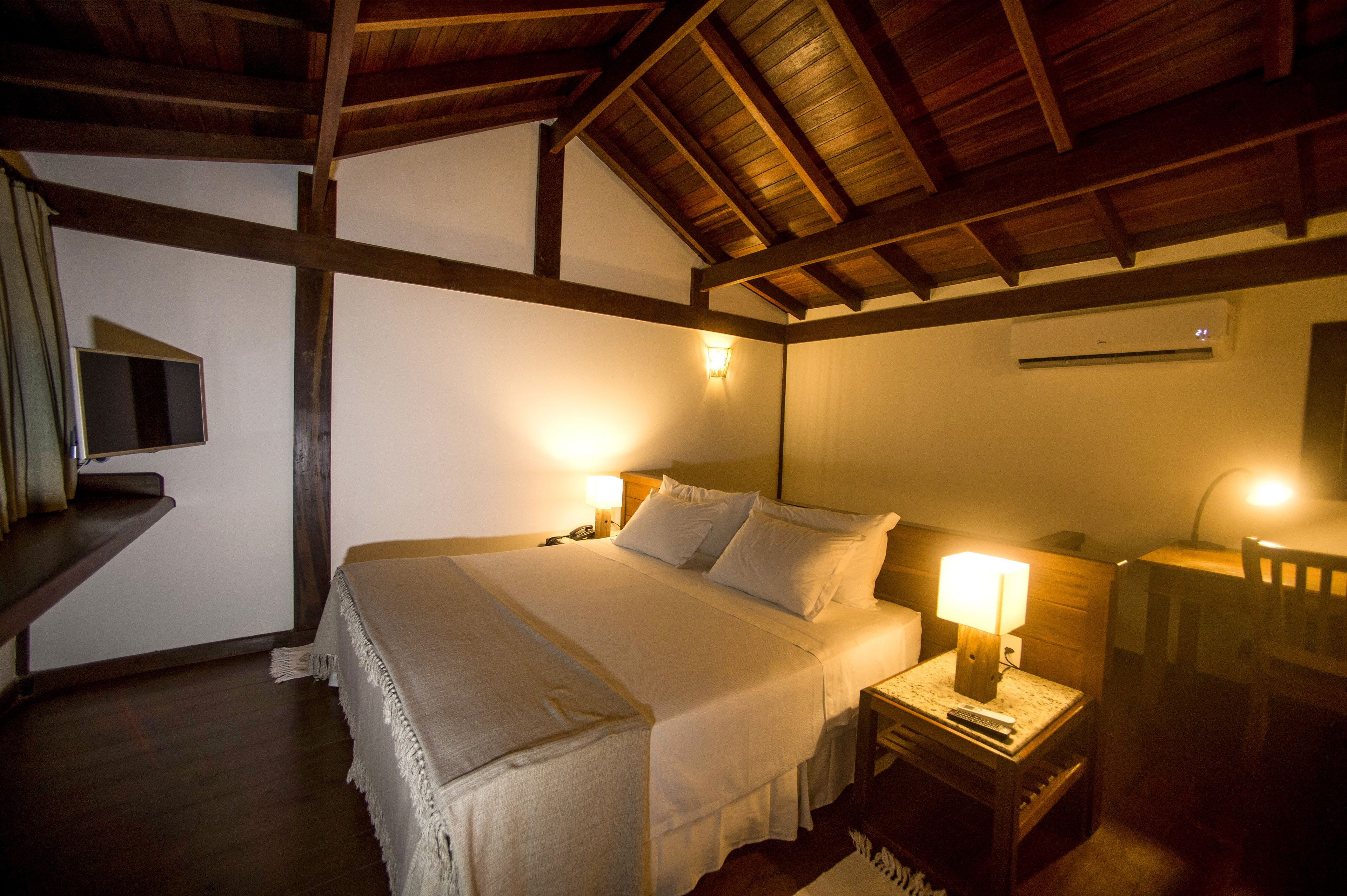 Une chambre avec un plafond en bois, un lit recouvert de draps blancs, deux lampes de chevet, une table de chevet et un climatiseur fixé au mur.