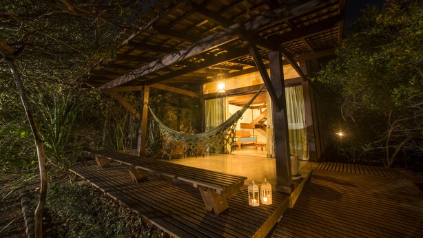 A wooden deck with a hammock and benches, leading to a building with a glass door and curtains.