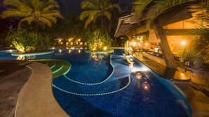 A nighttime swimming pool area with palm trees, lounge chairs, and a well-lit bar area.
