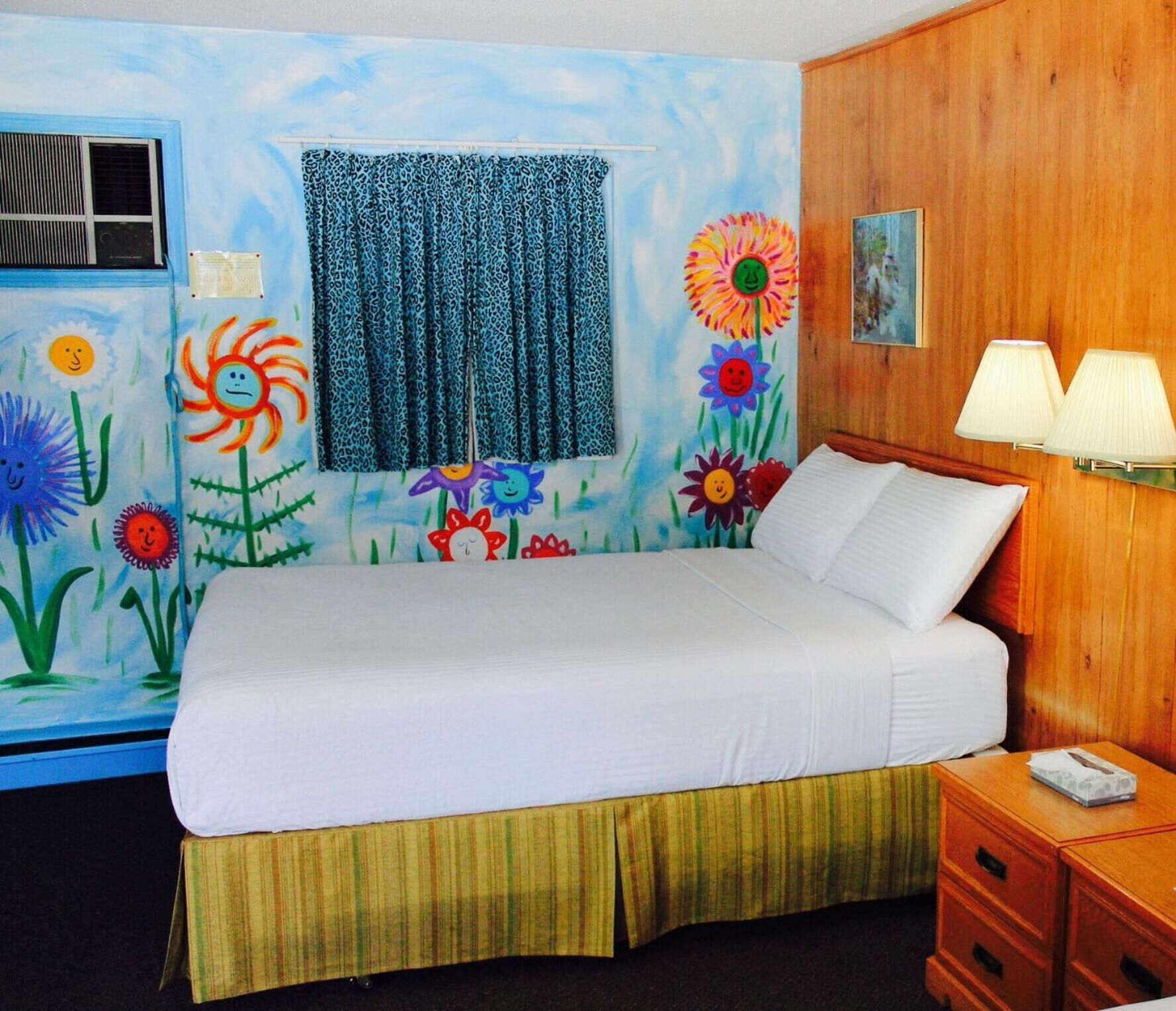 Un dormitorio con cama, ventana con cortina, pared con un mural floral, mesita de noche con lámpara y cabecera de madera.