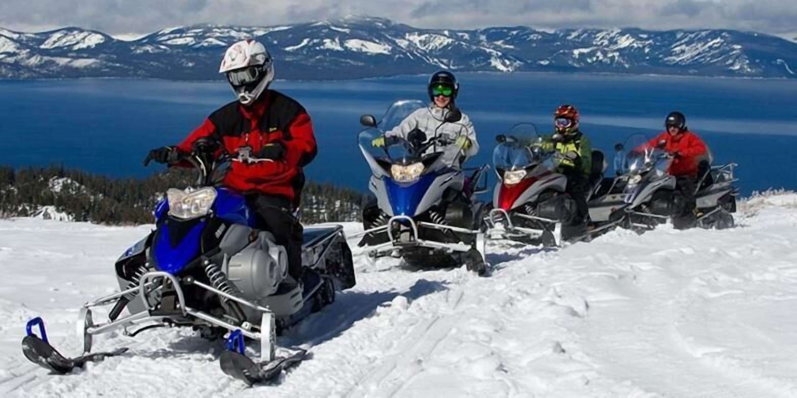 Cuatro motos de nieve en un paisaje nevado con un lago y montañas al fondo.