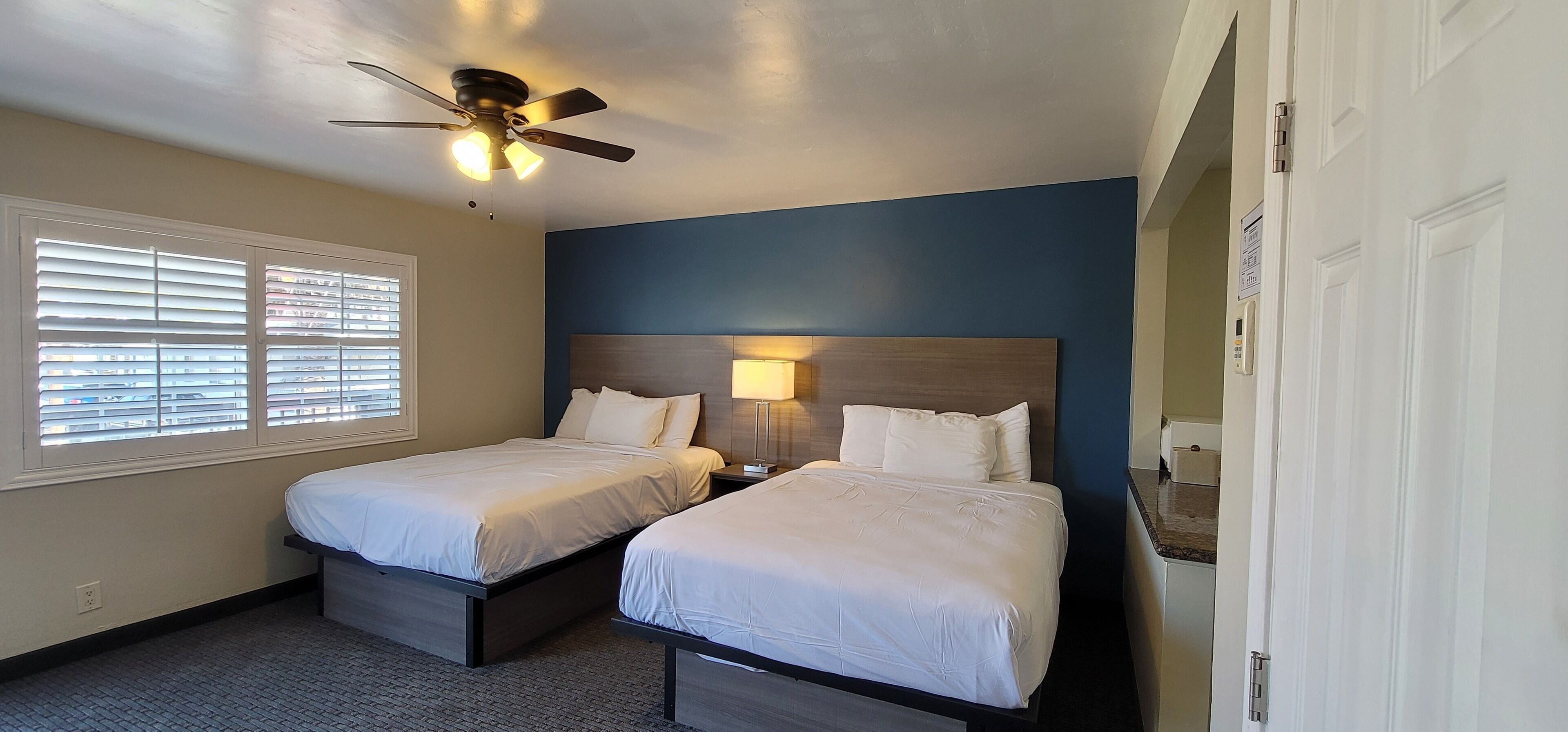 A hotel room with two beds, a ceiling fan, a nightstand, a lamp, and a window with blinds.