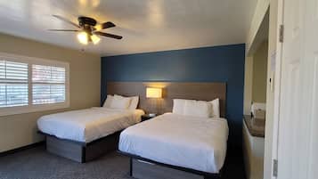 A hotel room with two beds, a ceiling fan, a nightstand, a lamp, and a window with blinds.
