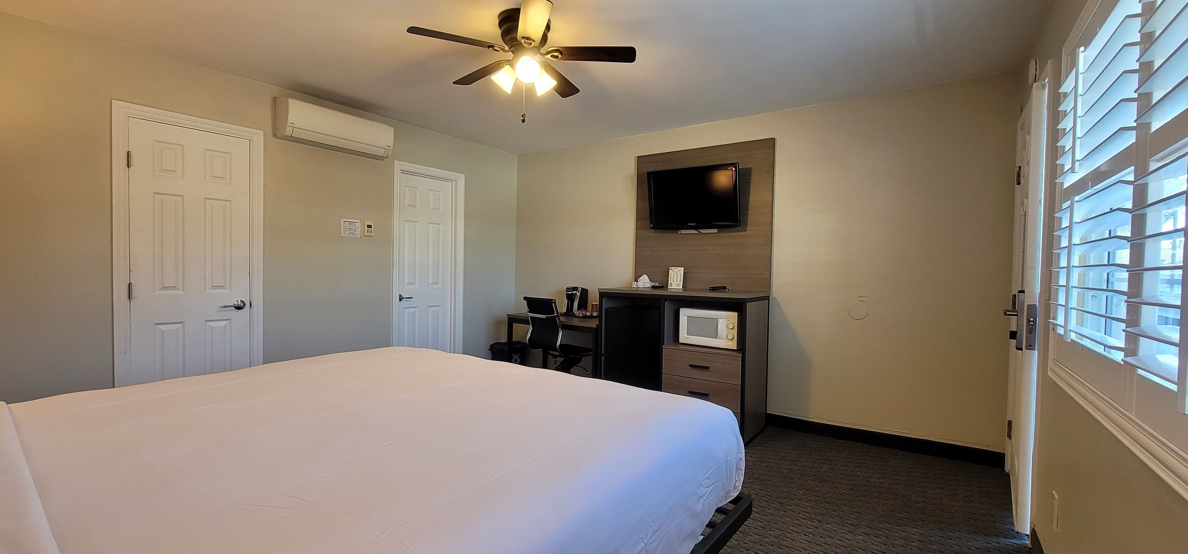 Una habitación de hotel con cama, escritorio con computadora, televisor montado en la pared y ventilador de techo.