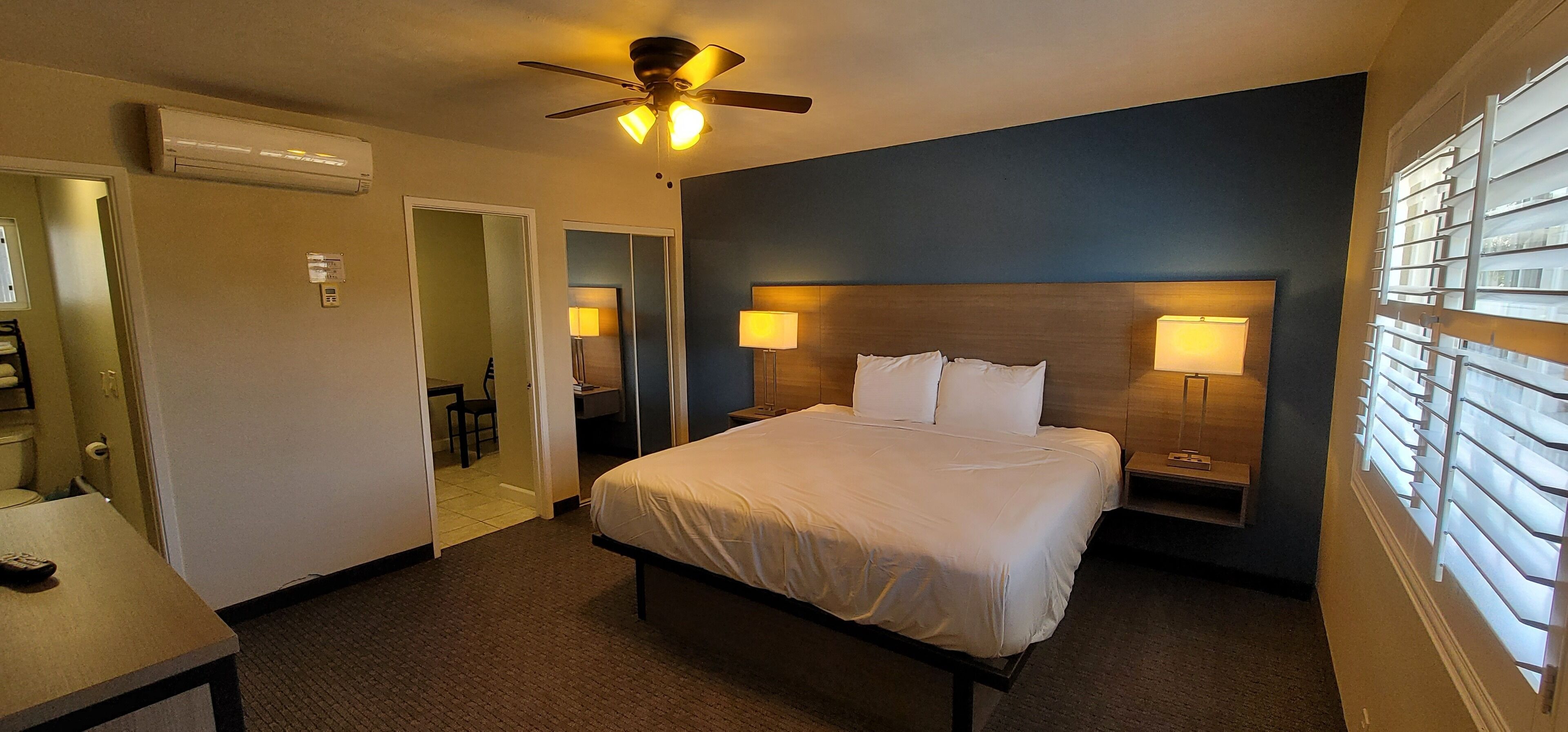 A hotel room with a bed, bedside lamps, a ceiling fan, an air conditioning unit, and a window with blinds.