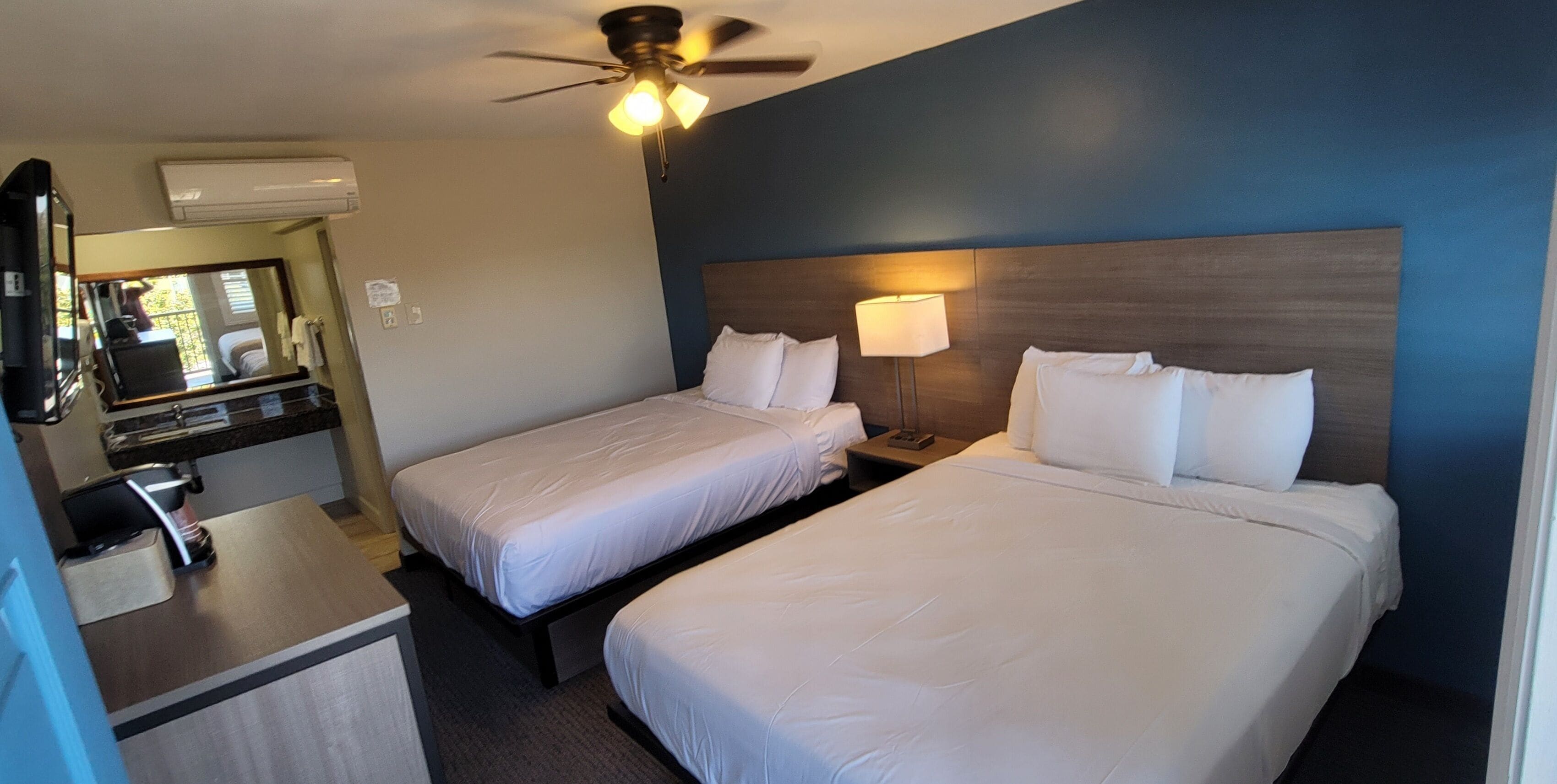 A hotel room with two beds, a desk with a coffee machine, a mirror, and a ceiling fan.