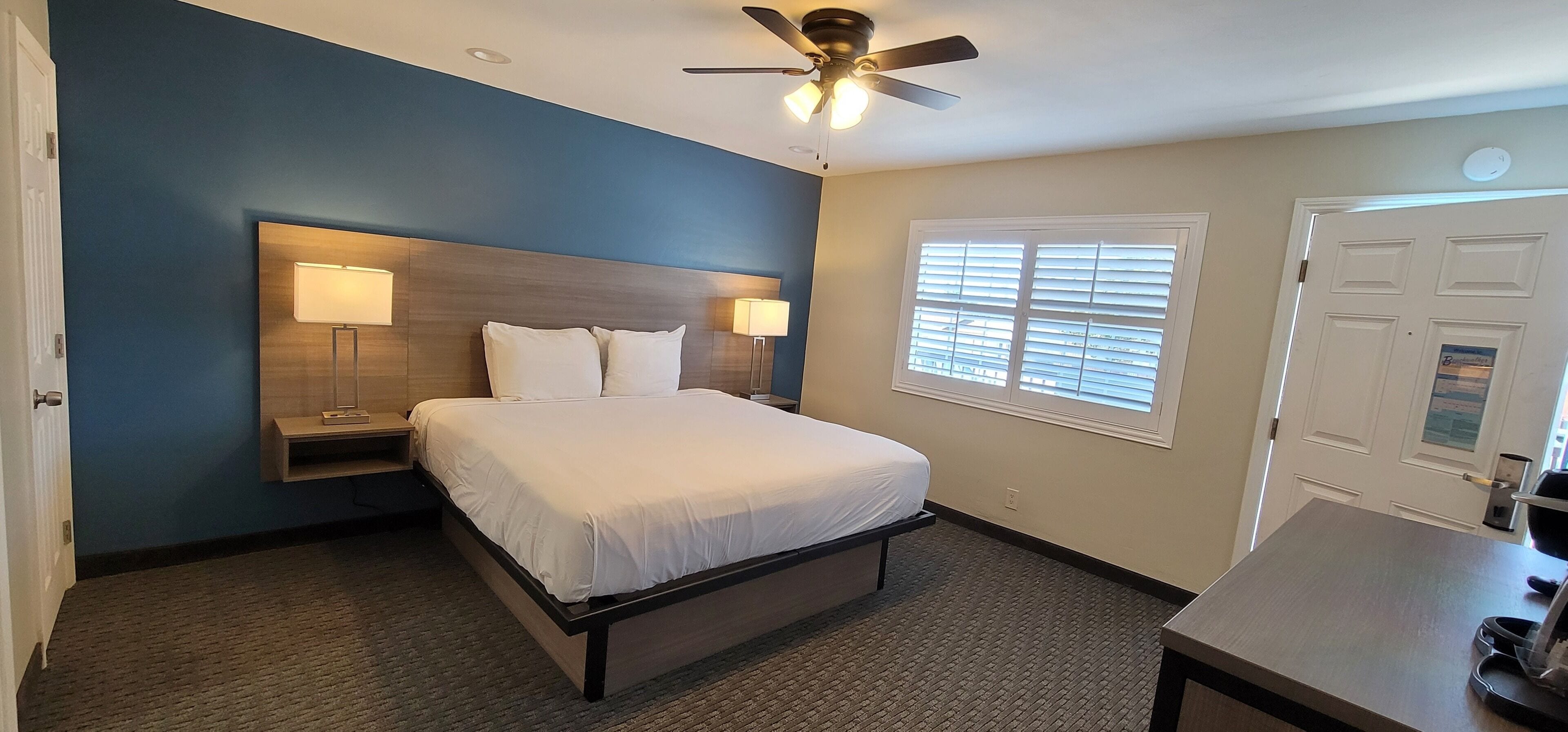 A bedroom with a bed, bedside lamps, a ceiling fan, a window with blinds, and a door.