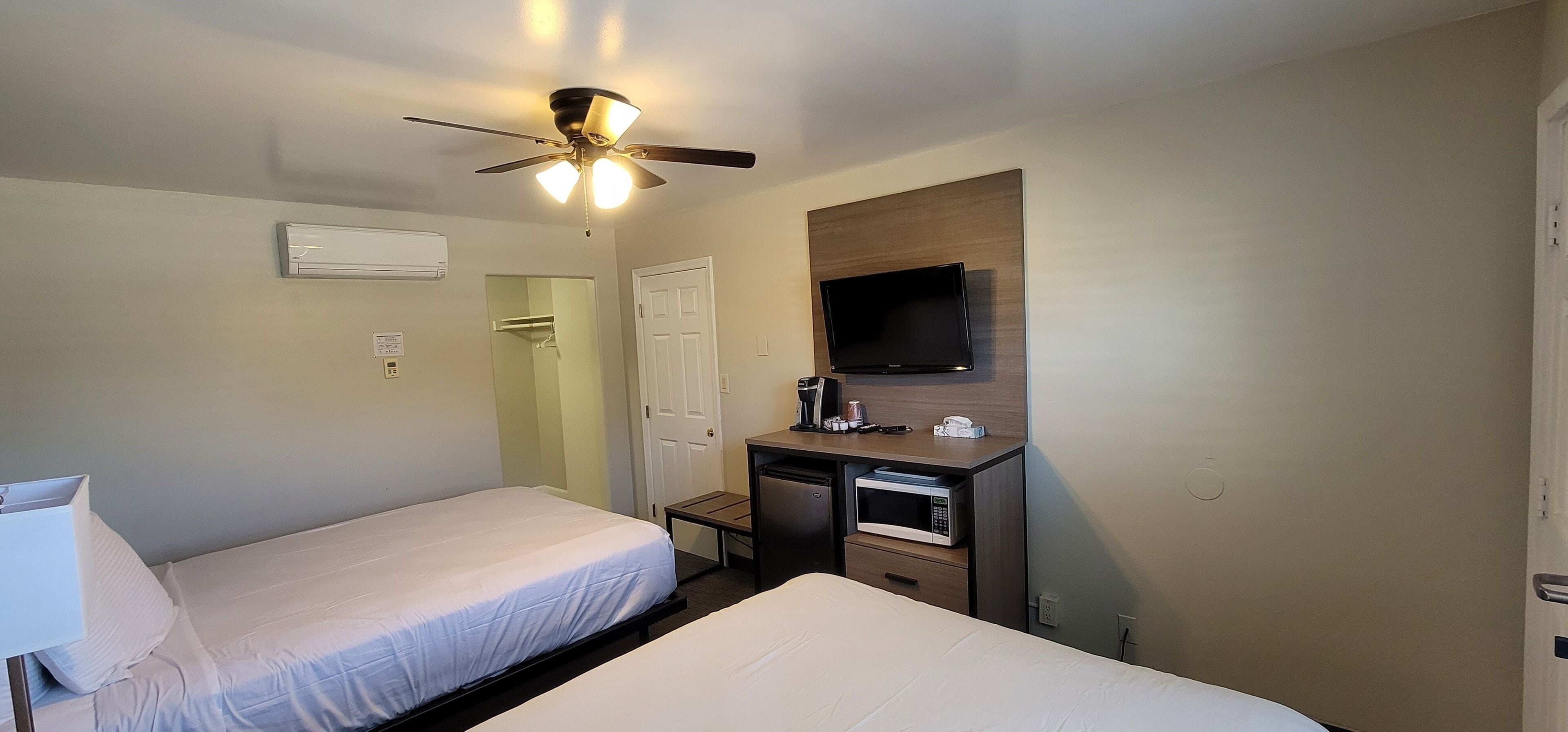 Habitación de hotel con dos camas individuales, ventilador de techo, televisor montado, escritorio con microondas y puerta que da a un baño.