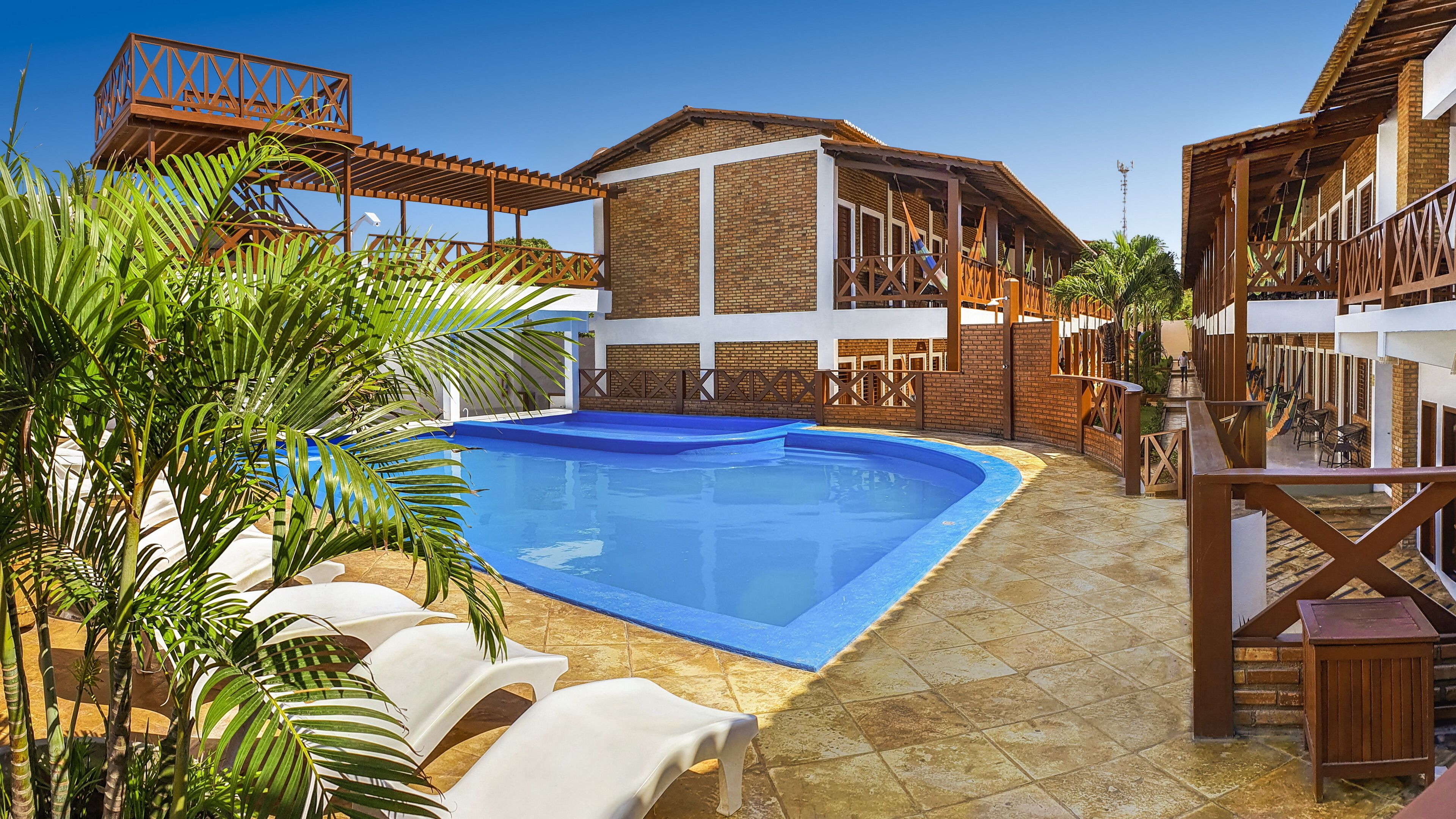 Una piscina con sillones blancos, un edificio de ladrillo con balcón y una pérgola de madera.