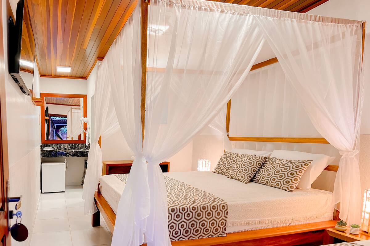 A bedroom with a four-poster canopy bed, a bedside table with a lamp, and a small potted plant.
