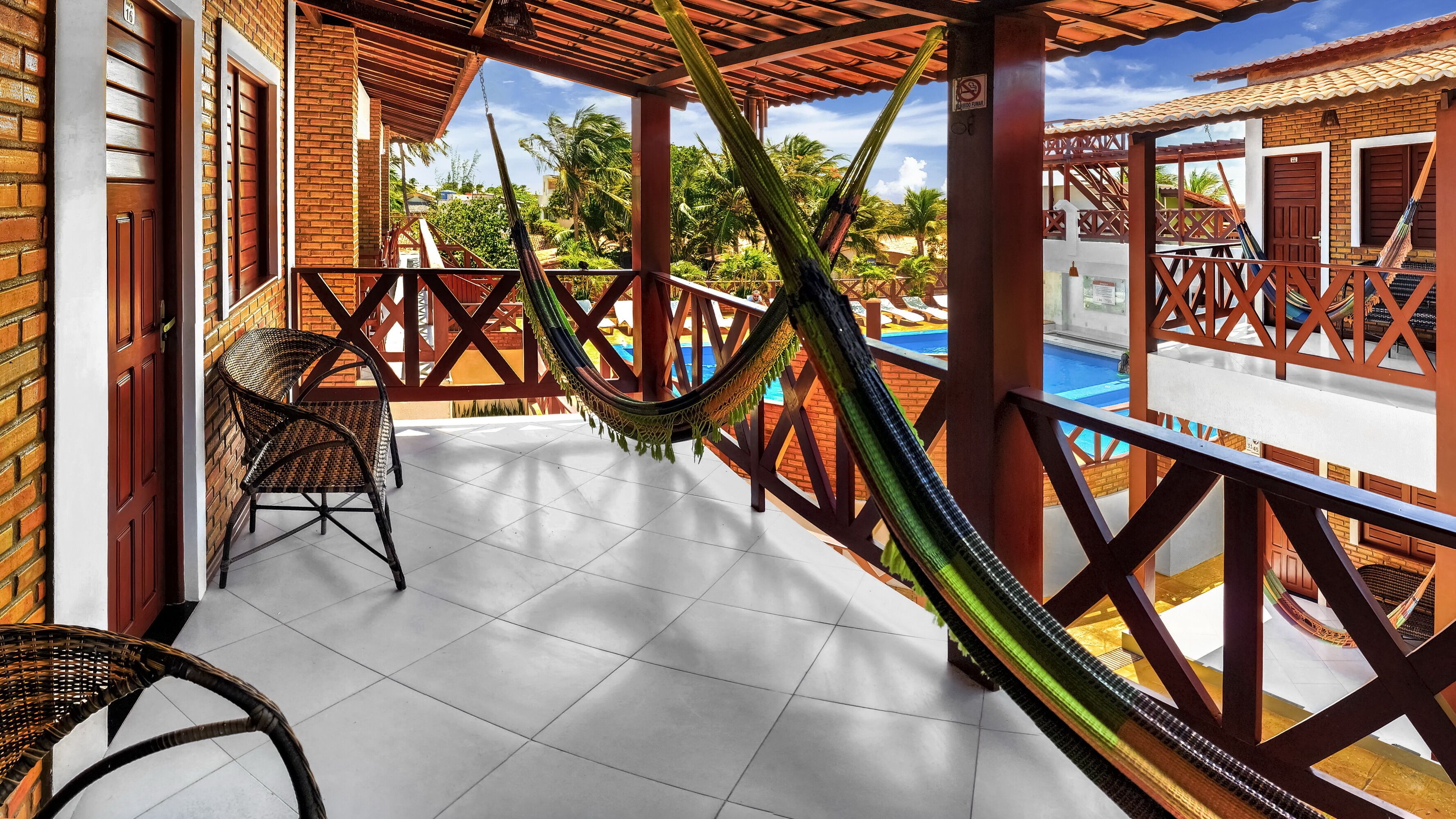 Un balcón techado con una hamaca, piscina y vistas a palmeras.