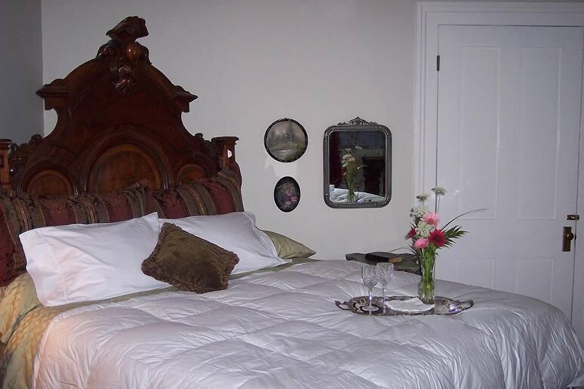 Ein ordentlich bezogenes Bett mit weißen Leinen, einem hölzernen Kopfteil und einem Nachttisch mit Blumenvase und Tablett mit Gläsern.