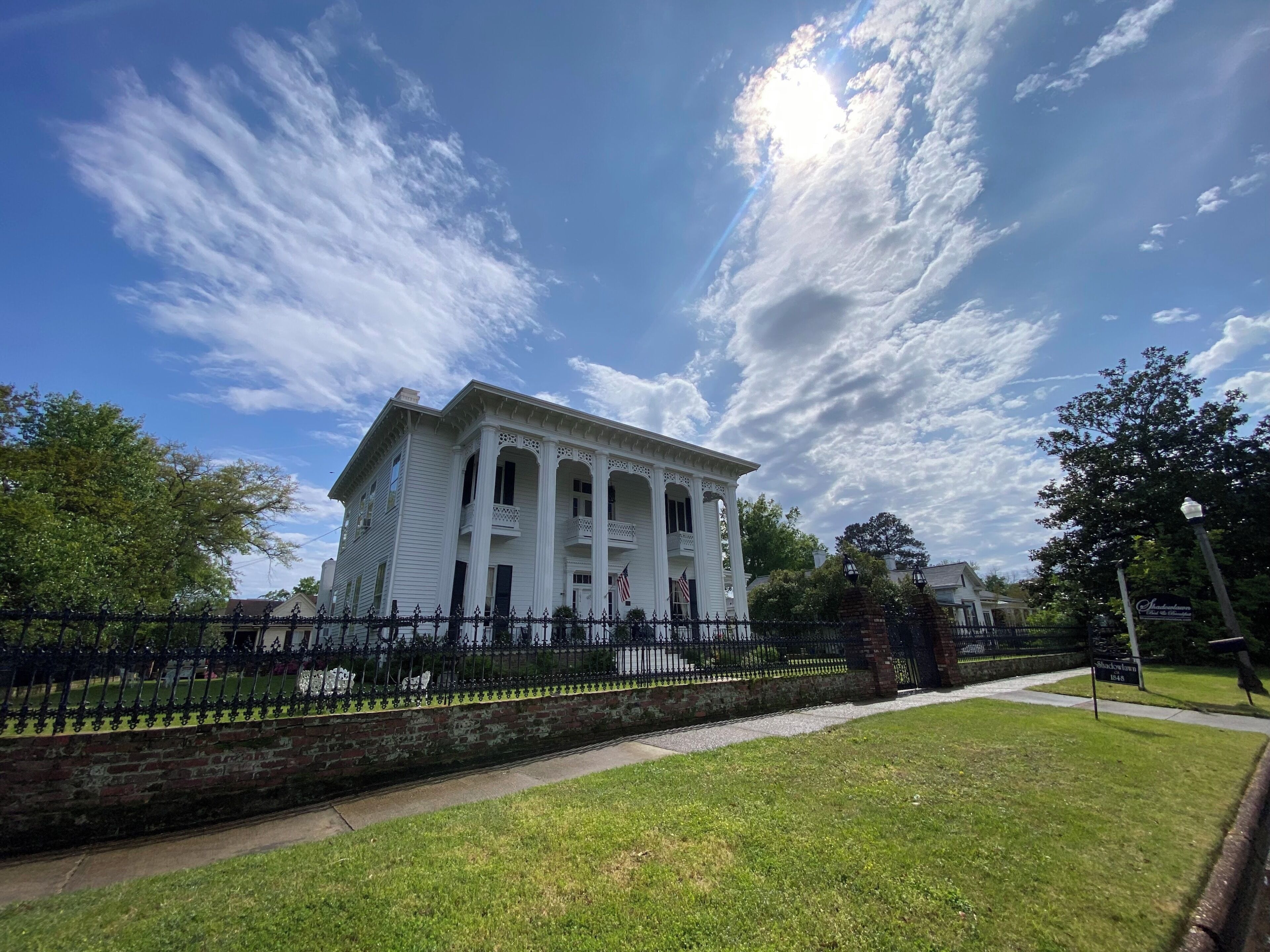 A historic white building with columns, a brick wall, and a black iron fence.