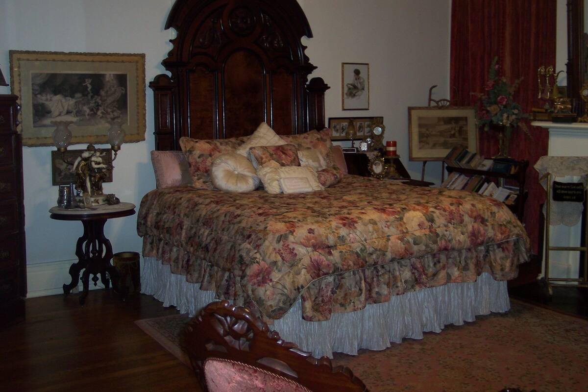 Ein traditionell eingerichtetes Schlafzimmer mit einem großen Bett, einem Holzkopfende und floralem Bettbezug. An den Wänden befinden sich mehrere gerahmte Bilder und ein kleiner Tisch mit einer Lampe.