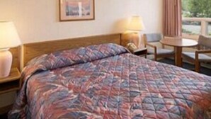 A hotel room with a bed, a chair, a table, a lamp, and a window with a view of greenery. - Aspen Inn (Grand Marais)