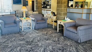 Un hall d’hôtel avec des fauteuils gris, une table basse, une cheminée et, au fond, un coin cuisine.