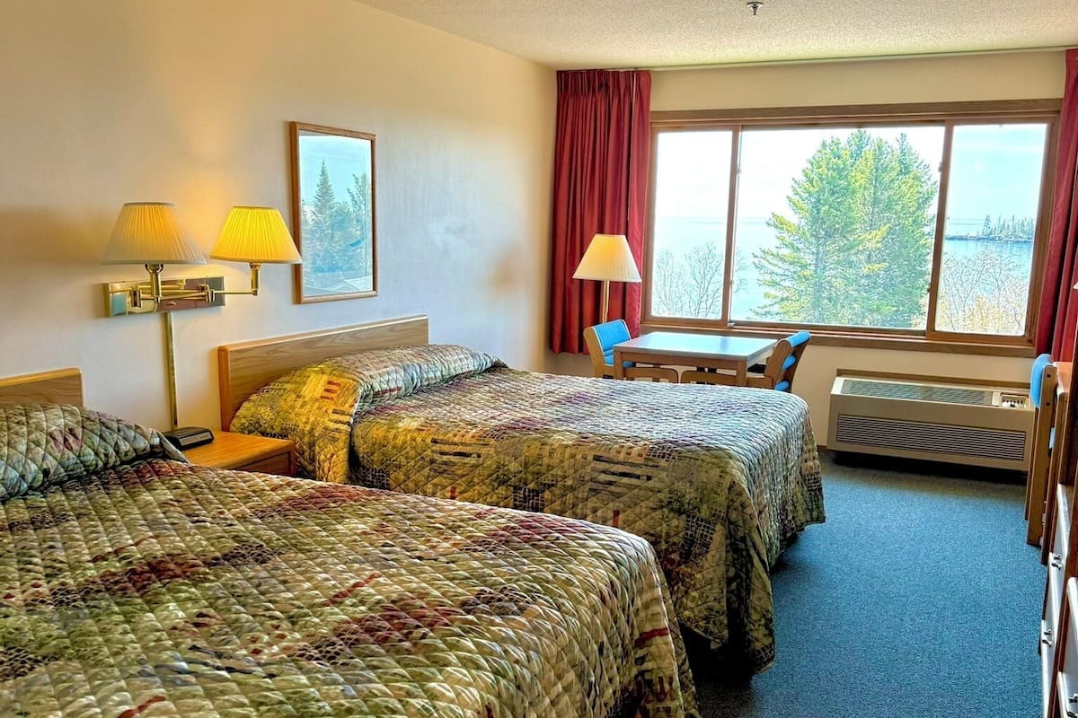 Habitación de hotel con dos camas, una mesa, sillas, un ventanal y una lámpara.