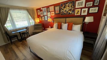 A hotel room with a large bed, a desk, and a chair. The room has a red wall with framed pictures and a clock. There is a window with curtains.