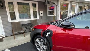 Ein rotes Auto ist an einer Ladestation vor einem Gebäude geparkt, an dem ein Schild mit der Aufschrift 'OPEN' hängt.
