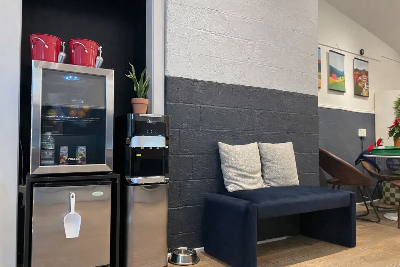 A compact living space with a kitchenette, a water dispenser, a small fridge, a seating area with cushions, and a dining table with chairs.