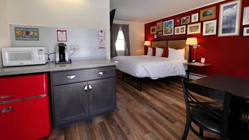 A compact hotel room with a kitchenette, a bed with white bedding, a red wall with framed pictures, and a wooden floor.