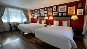 Ein Hotelzimmer mit zwei Betten, einem Schreibtisch und einem Stuhl. Der Raum hat Holzboden und rote Wände mit gerahmten Bildern.