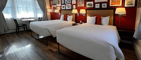 Habitación de hotel con dos camas, un escritorio y una silla. El suelo es de madera y las paredes son rojas con cuadros enmarcados.
