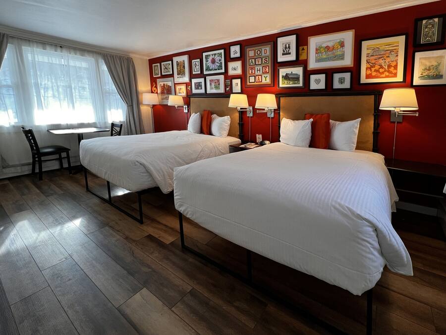 A hotel room with two beds, a desk, and a chair. The room has a wooden floor and red walls with framed pictures.