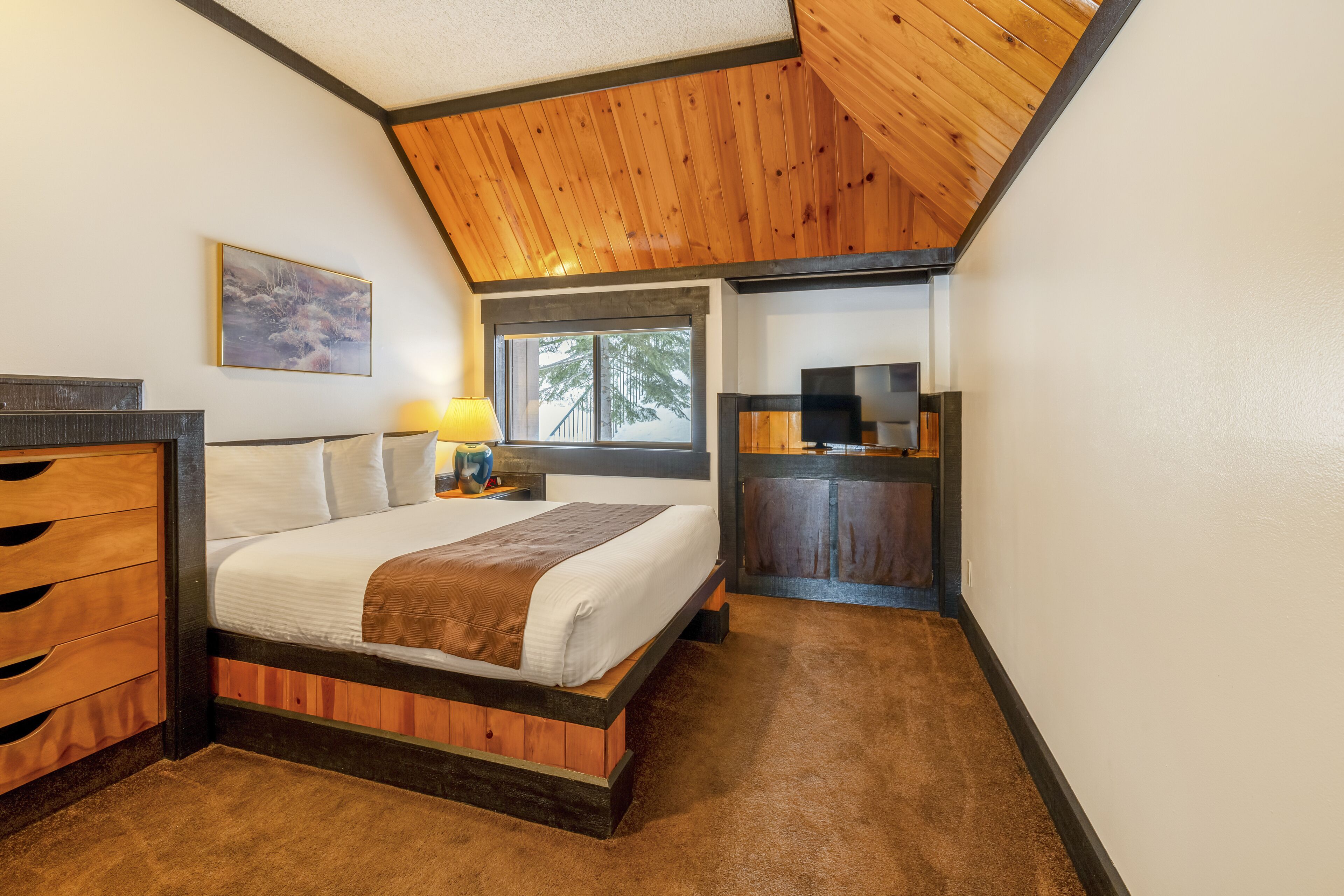 A bedroom with a wooden bed frame, a desk with a computer, and a window with a view of trees.